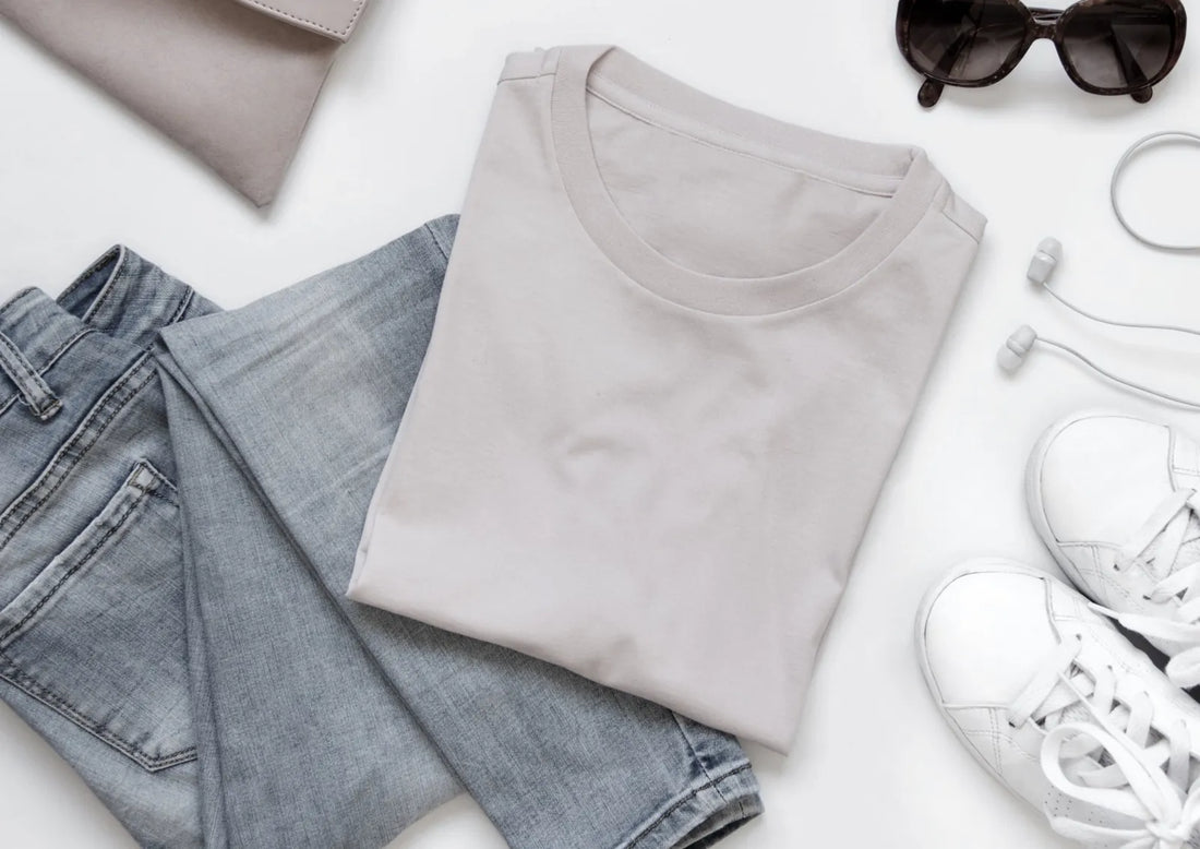 Capi basic del guardaroba disposti in flat lay: t-shirt grigio perla piegata, jeans chiari, sneakers bianche e occhiali da sole